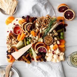 Overhead shot of a colorful charcuterie cheese board with assorted cheeses, crackers, dried apricots and figs, blood orange slices, walnuts, pistachios, olives, cornichons, grainy mustard and honey, styled with rosemary and a glass of white wine