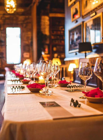 Cozy candlelit restaurant interior with a long white-clothed table set for dinner—rows of wine glasses, bowls with red napkins, eucalyptus sprigs and framed artwork on the walls.