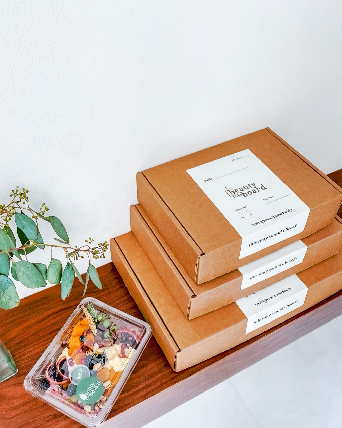 Stacked kraft delivery boxes with white labels next to a clear plastic grazing box of colorful cheeses, fruits, nuts and edible flowers on a wooden table with a sprig of eucalyptus against a white wall.