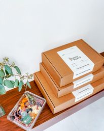Stacked kraft delivery boxes with white labels next to a clear plastic grazing box of colorful cheeses, fruits, nuts and edible flowers on a wooden table with a sprig of eucalyptus against a white wall.