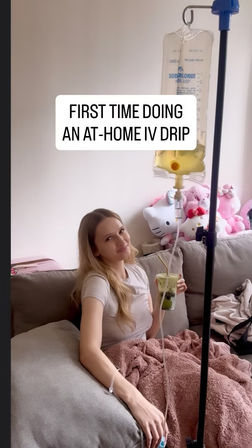 Woman relaxing on a living-room couch receiving an at-home IV drip from a hanging saline bag, sipping an iced matcha drink and wrapped in a pink blanket with plush toys in the background — cozy home wellness scene.