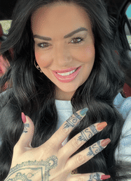 Close-up car selfie of a smiling woman with long black hair, glossy pink lips, multicolored almond nails and intricate finger and hand tattoos.