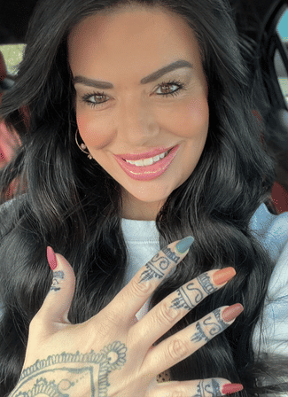 Close-up car selfie of a smiling woman with long black hair, glossy pink lips, multicolored almond nails and intricate finger and hand tattoos.