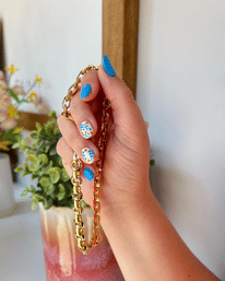 Close-up of a hand with bright blue polish and white floral nail art holding a chunky gold chain above a potted succulent