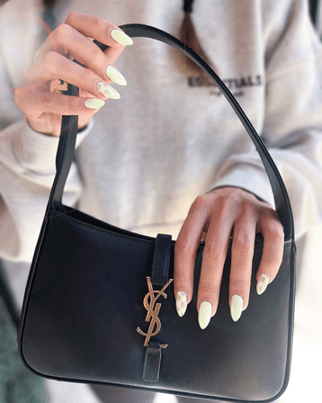Close-up of hands with pastel neon-green almond-shaped nail art holding a black leather shoulder bag with gold metal logo accent against a light sweatshirt — casual street-style accessory detail.