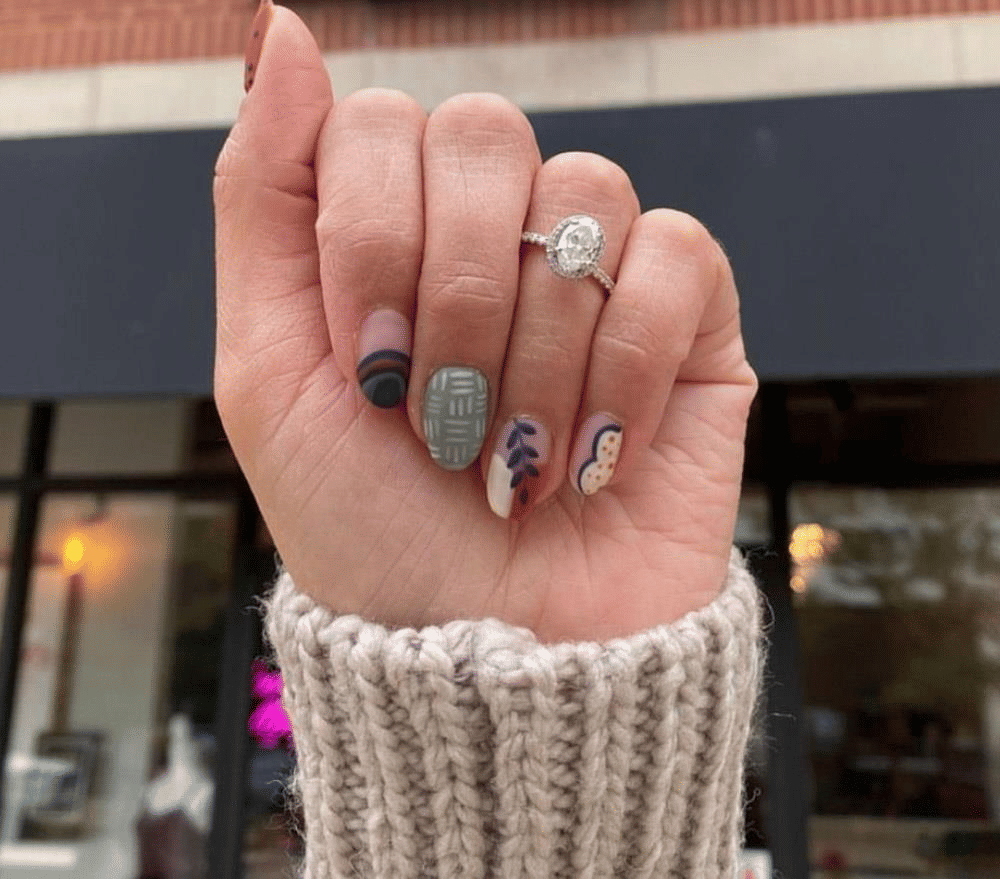 Close-up of a hand wearing an oval halo engagement ring and chunky knit cuff, showing fall-inspired manicure and nail art—matte sage plaid, abstract dots and leaf motifs—held up in front of a blurred storefront.