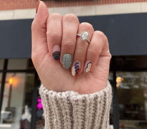 Close-up of a hand wearing an oval halo engagement ring and chunky knit cuff, showing fall-inspired manicure and nail art—matte sage plaid, abstract dots and leaf motifs—held up in front of a blurred storefront.
