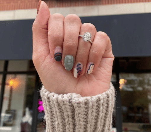 Close-up of a hand wearing an oval halo engagement ring and chunky knit cuff, showing fall-inspired manicure and nail art—matte sage plaid, abstract dots and leaf motifs—held up in front of a blurred storefront.