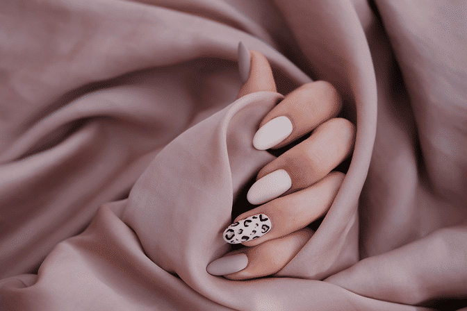 Close-up of a hand with nude matte almond-shaped nails and a leopard-print accent nail resting on blush satin fabric.