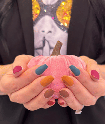 Close-up of hands with matte multicolor fall manicure (teal, mustard, brown, magenta) holding a small pink velvet pumpkin, seasonal autumn manicure close-up