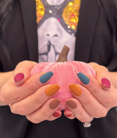 Close-up of hands with matte multicolor fall manicure (teal, mustard, brown, magenta) holding a small pink velvet pumpkin, seasonal autumn manicure close-up