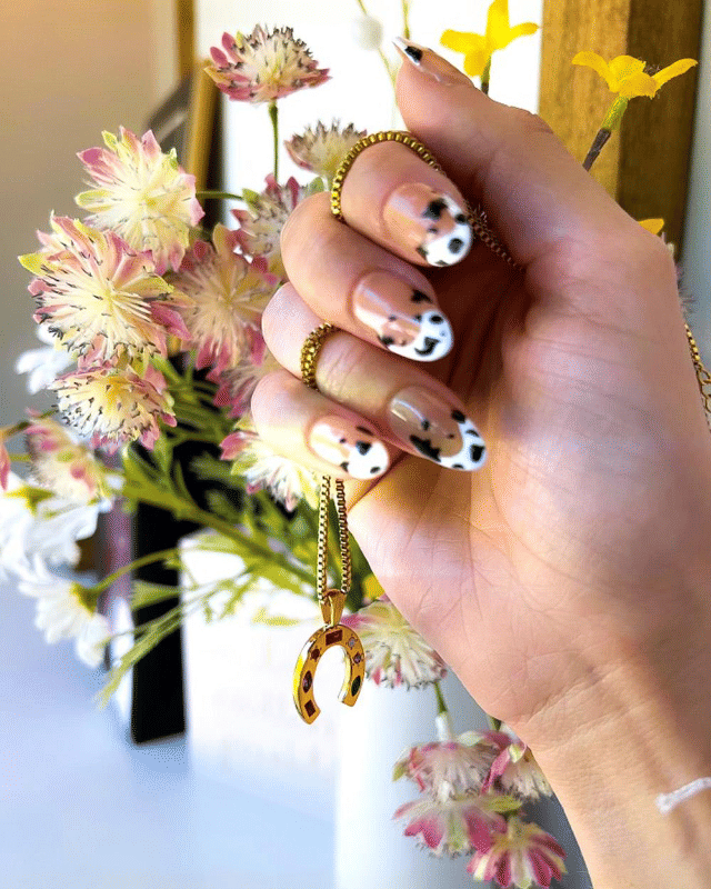 Close-up of hand with nude manicure and black-and-white cow-print nail art holding a gold horseshoe pendant on a chain against a pink-and-white floral bouquet background.
