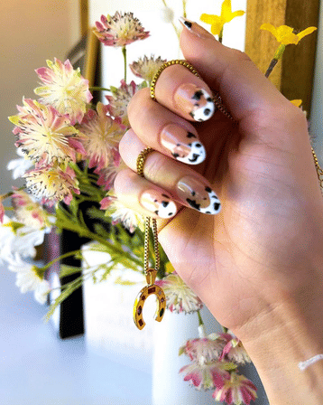 Close-up of hand with nude manicure and black-and-white cow-print nail art holding a gold horseshoe pendant on a chain against a pink-and-white floral bouquet background.