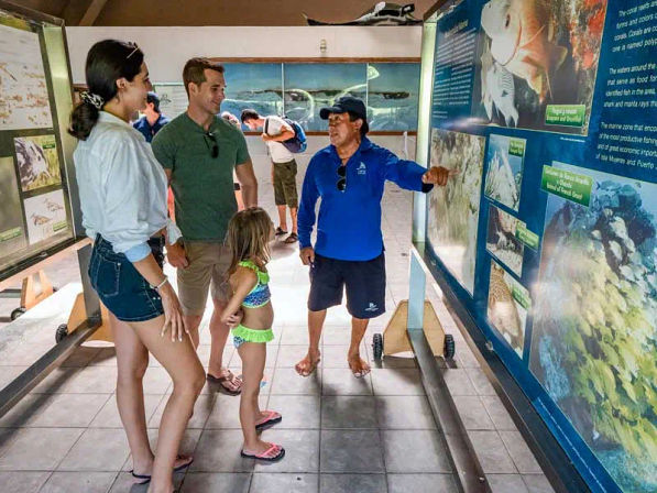 Family with young child listening to a guide pointing at large interpretive panels about coral reefs and fish in a coastal marine exhibit