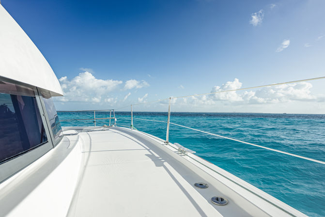 Sunlit yacht deck gliding across turquoise tropical sea under a bright blue sky with fluffy clouds on the horizon.