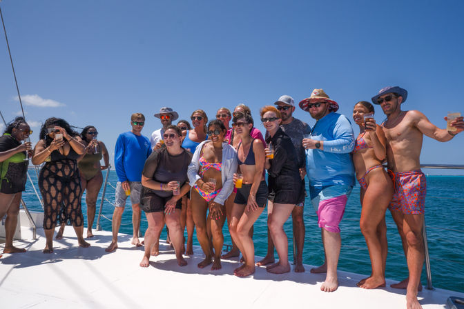 Group of adults on a catamaran deck in clear tropical blue ocean, wearing swimsuits and sun hats, holding drinks and posing for a sunny boat party
