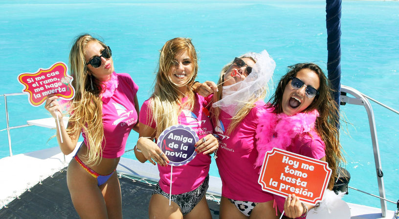 Four friends in matching pink shirts on a catamaran in turquoise Cancún waters, smiling and holding playful Spanish bachelorette party signs.