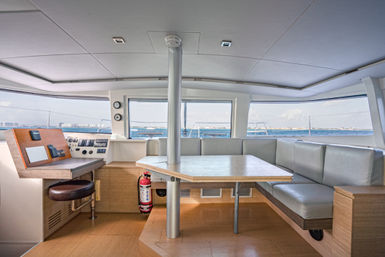 Bright modern catamaran salon and helm with wooden dinette table, L-shaped gray cushioned seating, navigation console and stool, and panoramic ocean view through wraparound windows.