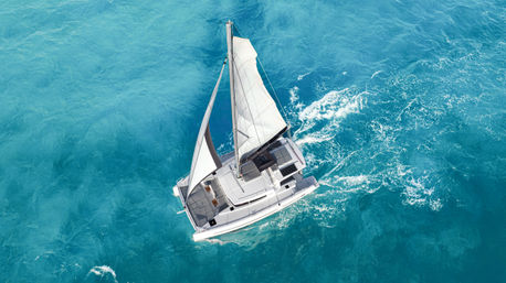 Aerial view of a white catamaran sailboat gliding across turquoise ocean water, its white sails up and a foamy wake trailing behind.