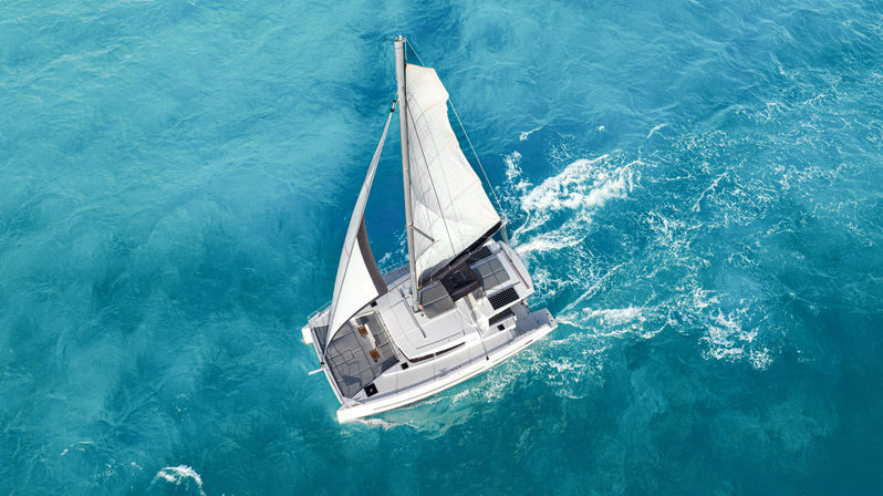 Aerial view of a white catamaran sailboat gliding across turquoise ocean water, its white sails up and a foamy wake trailing behind.