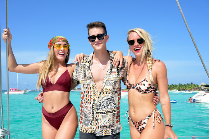 Three smiling friends in sunglasses and beachwear posing on a sailboat over turquoise tropical waters with palm-lined island and other boats in the sunny background.