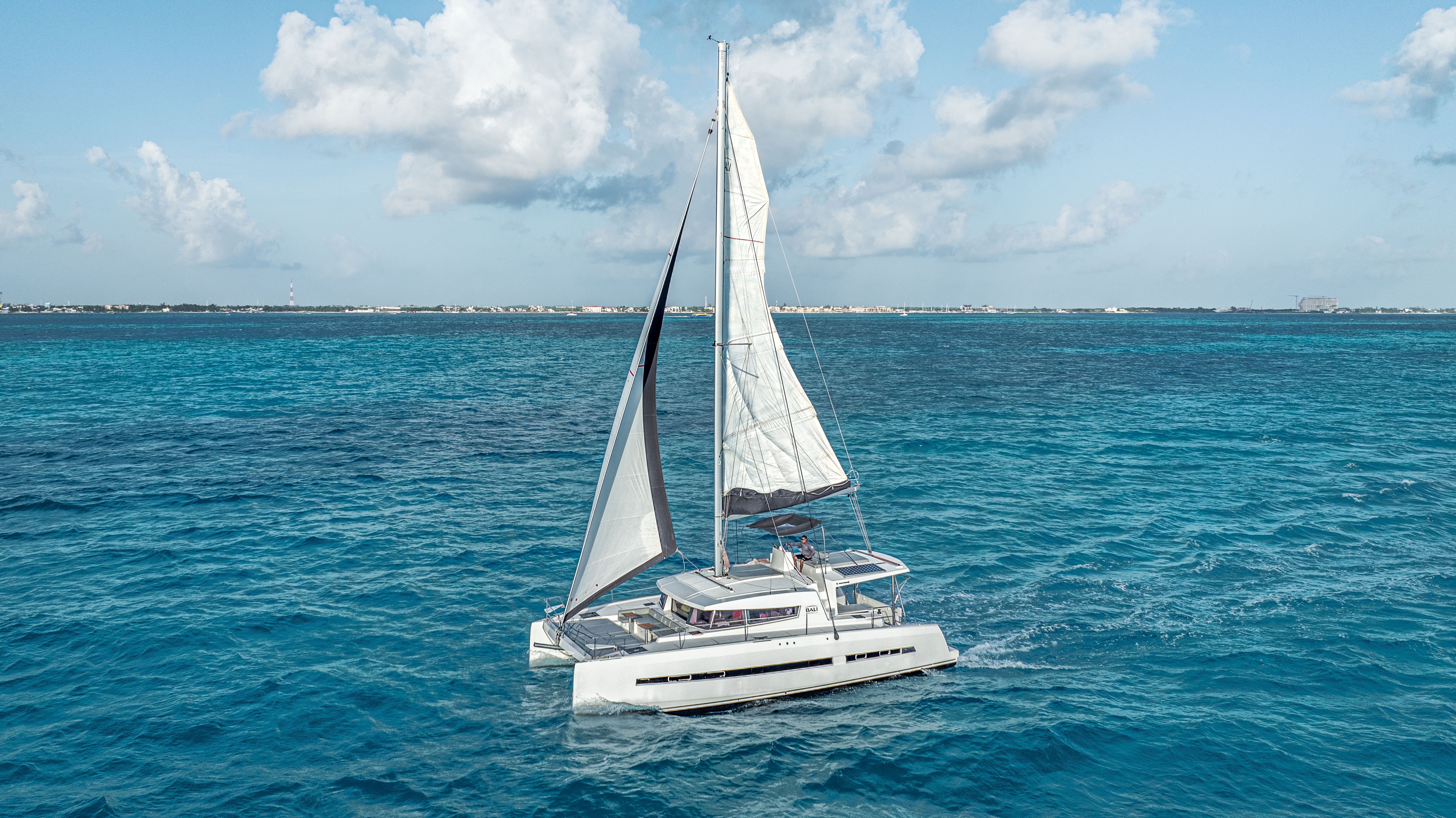 White catamaran sailboat cruising on vivid turquoise water under a sunny, partly cloudy sky with a low coastline visible on the horizon.