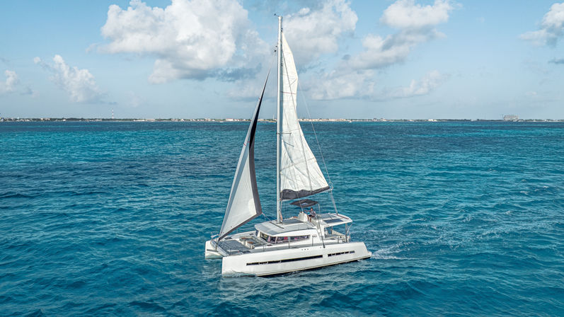 White catamaran sailboat cruising on vivid turquoise water under a sunny, partly cloudy sky with a low coastline visible on the horizon.