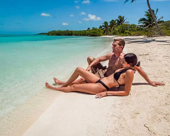 Smiling couple in swimsuits lounging on a white-sand tropical beach with clear turquoise water, palm trees and a bright blue sky.