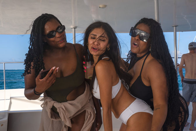 Three women in swimsuits and sunglasses laughing and posing on a sunlit yacht deck with deep blue ocean in the background, one holding a drink.