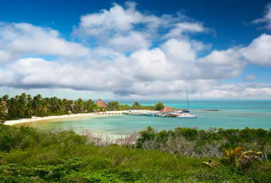 Turquoise tropical bay with white-sand beach and palm trees, a wooden pier with a moored sailboat, lush green foreground, and blue sky dotted with puffy clouds.