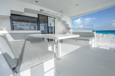 Sunlit white catamaran deck dining area with cushioned bench seats and a central table overlooking a turquoise sea and bright blue sky