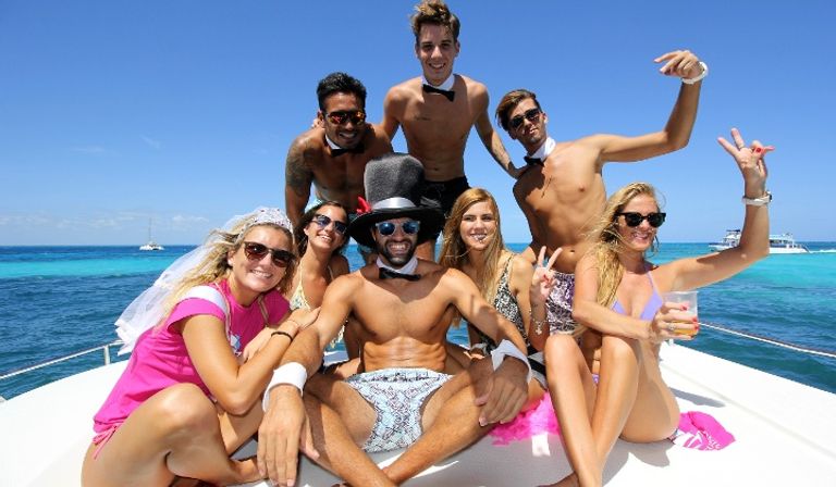 Group of friends on a yacht in turquoise tropical water, smiling in swimwear and playful costumes (top hat, veil, bow ties) during a sunny boat party