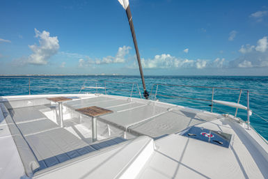Sun-drenched catamaran foredeck with cushioned lounge seating, two small wooden tables and railings overlooking turquoise tropical sea and blue sky with scattered clouds.