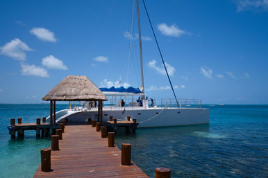 Sunny tropical wooden dock with a thatched palapa and a moored white catamaran on clear turquoise water under a bright blue sky — perfect island vacation scene.