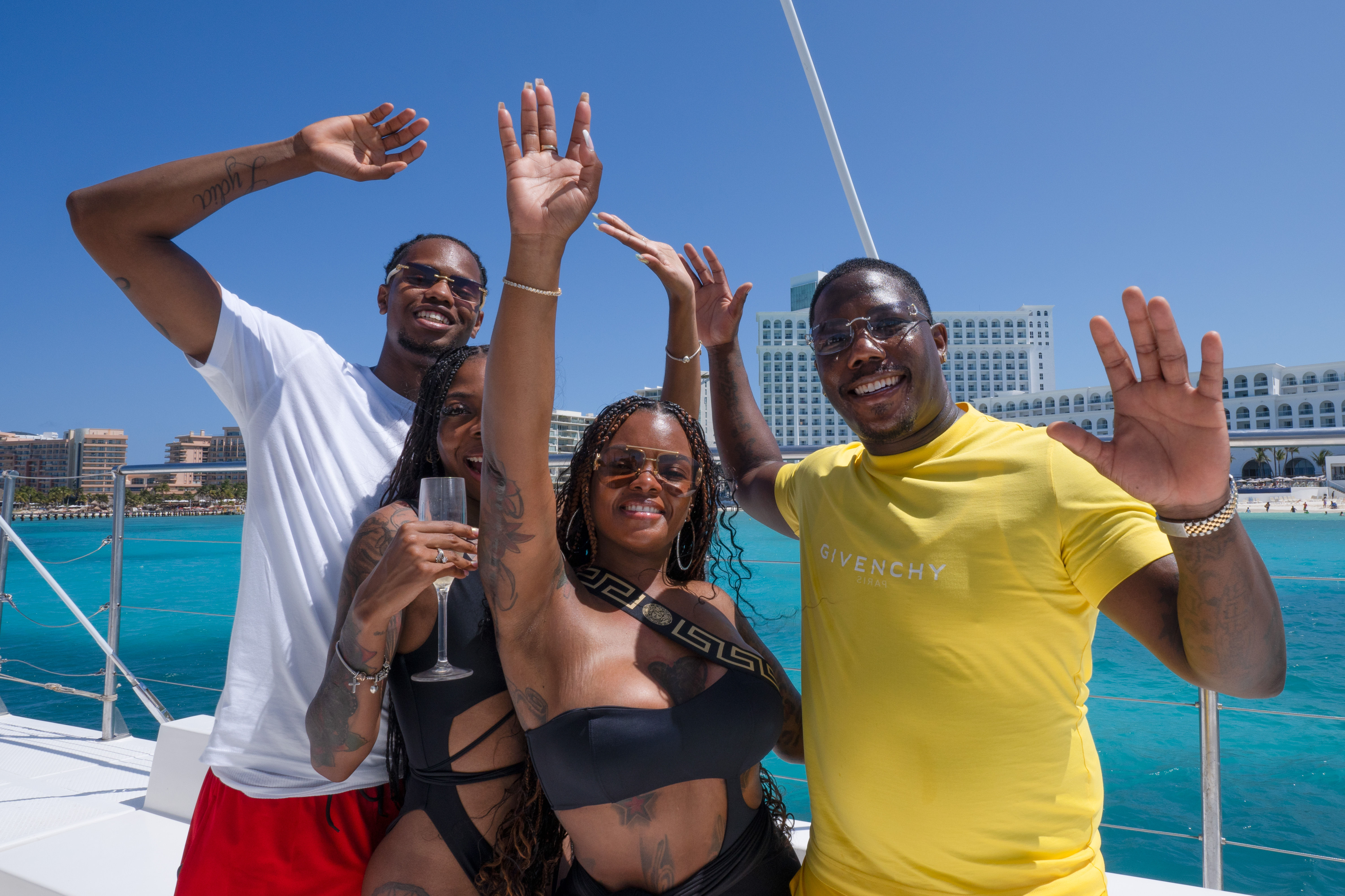 Four friends on a boat waving and smiling in swimwear and summer clothes, one holding a champagne flute, with bright turquoise ocean and a large beachfront resort under a clear blue sky.