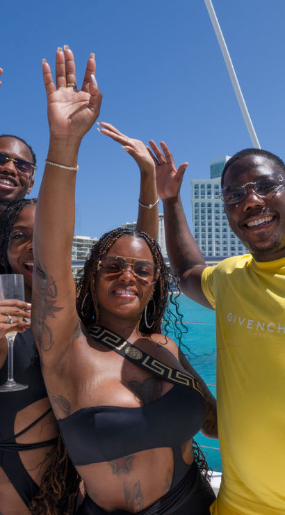 Four friends on a boat waving and smiling in swimwear and summer clothes, one holding a champagne flute, with bright turquoise ocean and a large beachfront resort under a clear blue sky.