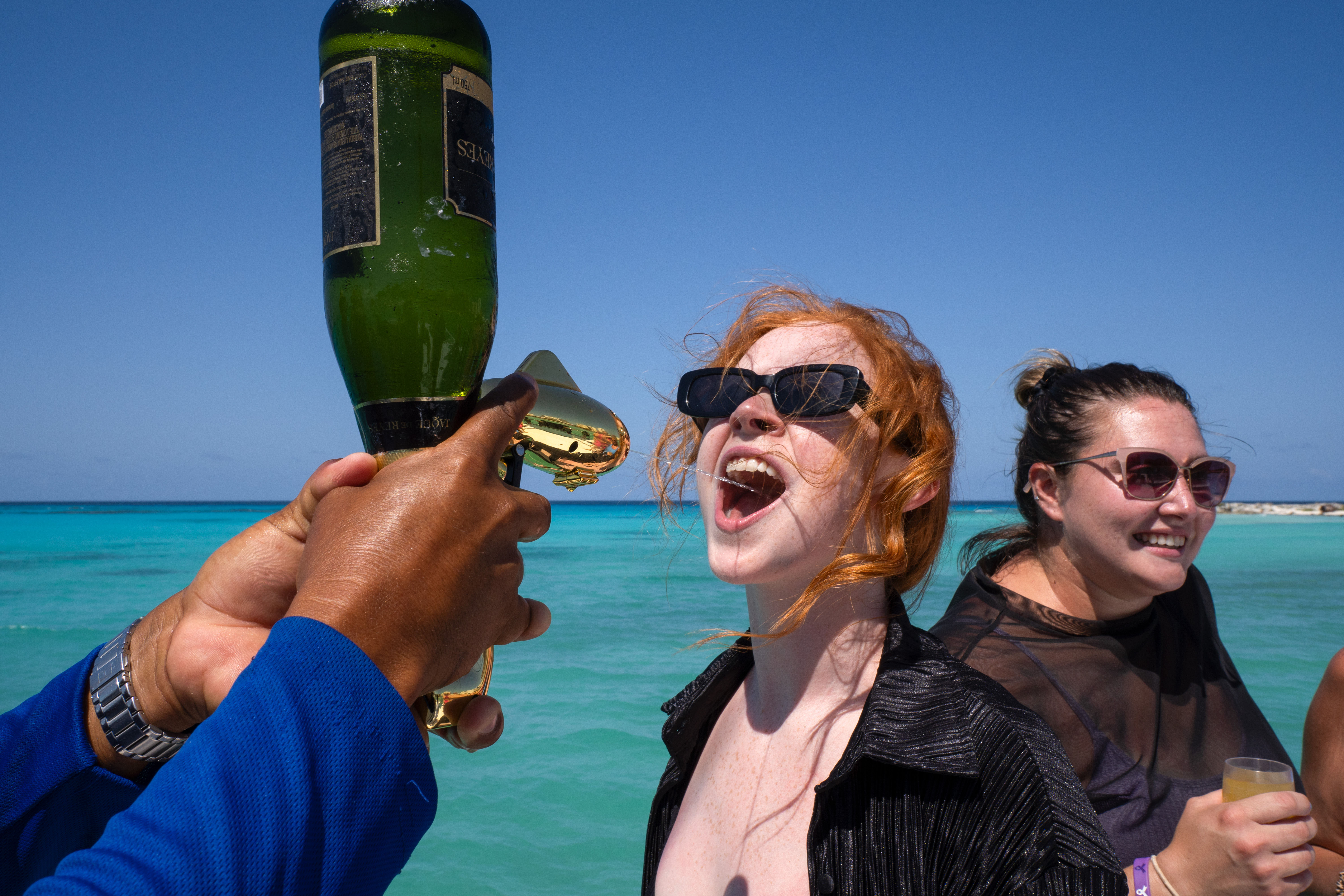 Two women in sunglasses on a boat during a tropical vacation — one laughing with her mouth open as a server pours champagne from a green bottle into it against turquoise water and a clear blue sky.