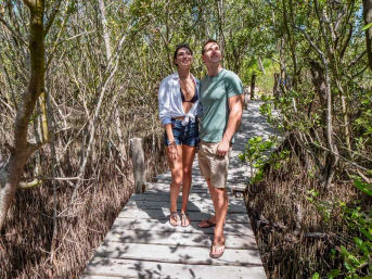 Man and woman standing on a sunny wooden boardwalk through dense coastal mangroves, smiling and looking up in casual summer clothes and sandals