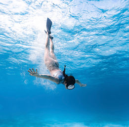 Snorkeler with fins diving beneath sunlit, crystal-clear tropical blue water, bubbles rising toward the rippled surface.
