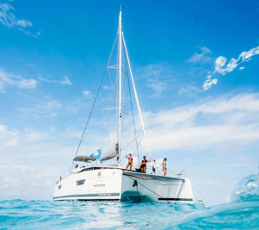 White catamaran on clear turquoise ocean under a bright blue sky, people relaxing on the bow enjoying a sunny day at sea.