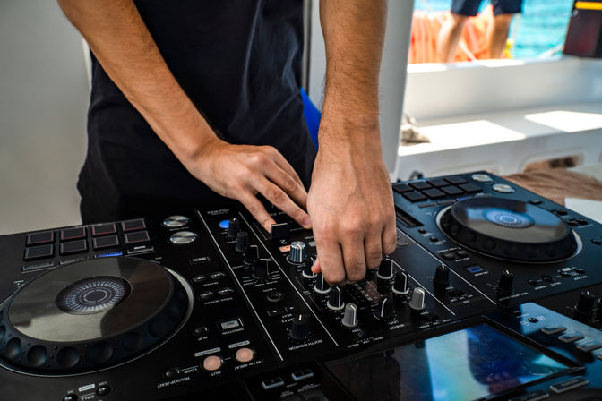 Close-up of DJ hands adjusting knobs on a professional mixer and controller aboard a boat with ocean in the background, seaside party vibe.