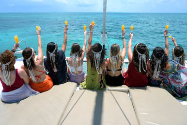 Back view of women wearing macramé headpieces on a catamaran, toasting with mimosas toward a turquoise tropical ocean during a sunny boat celebration.
