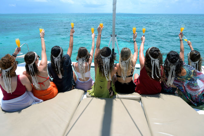 Back view of women wearing macramé headpieces on a catamaran, toasting with mimosas toward a turquoise tropical ocean during a sunny boat celebration.