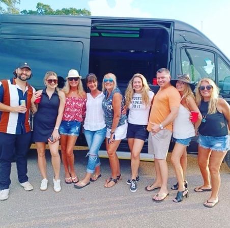 Nine adults in summer clothes smiling and posing in front of a black passenger van in a sunny parking lot, some holding drinks and red cups — casual road-trip or tailgate group photo.