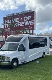 White shuttle bus parked on grass in front of a large red stadium billboard reading 'HOME OF THE KREWE', palm trees and cloudy sky