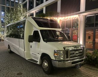 Sleek white shuttle minibus parked on a cobblestone driveway outside a downtown hotel entrance at night, with trees and marquee wrapped in warm string lights.