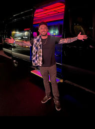 Smiling tattooed man in casual clothes stands with arms outstretched outside a black party van lit by pink and blue neon lights at night