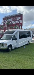 White charter shuttle bus parked on grass in front of a large red stadium sign with palm trees and a cloudy sky, game-day parking lot scene.