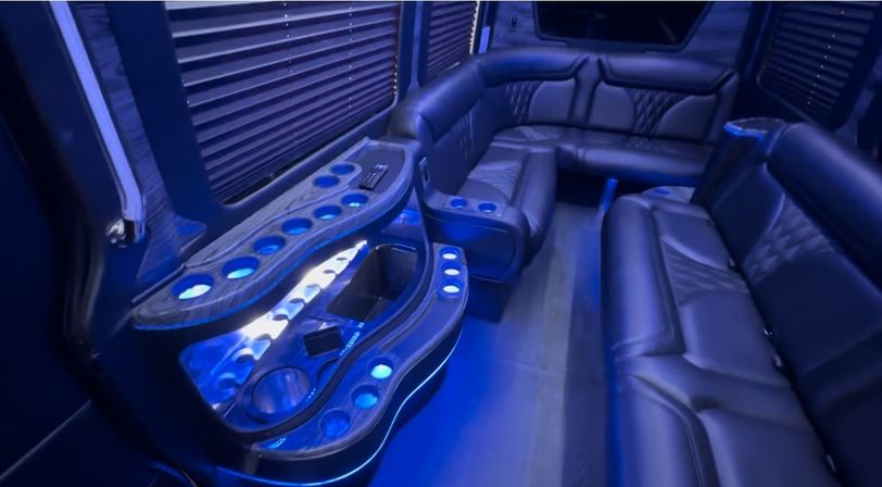 Sleek VIP party-bus interior bathed in blue LED light with wraparound black leather seating, illuminated cup holders and a built-in mini bar.