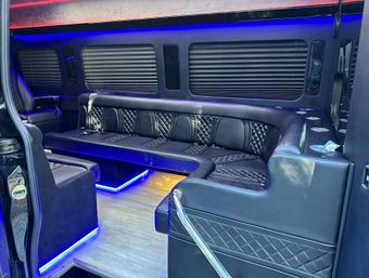 Luxurious black leather U-shaped seating inside a party van or limo with quilted upholstery, window blinds, cup holders and vibrant blue LED accent lighting under the seats and floor.